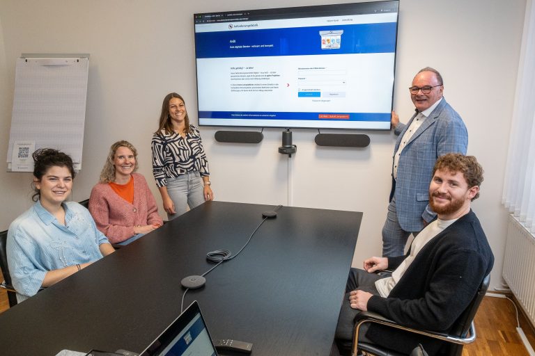 Präsentation Projekt "AnDi": Es sind fünf Personen um einen Tisch versammelt, im Hintergrund am Display ist das Lernwerk der Anforderungsfabrik zu sehen