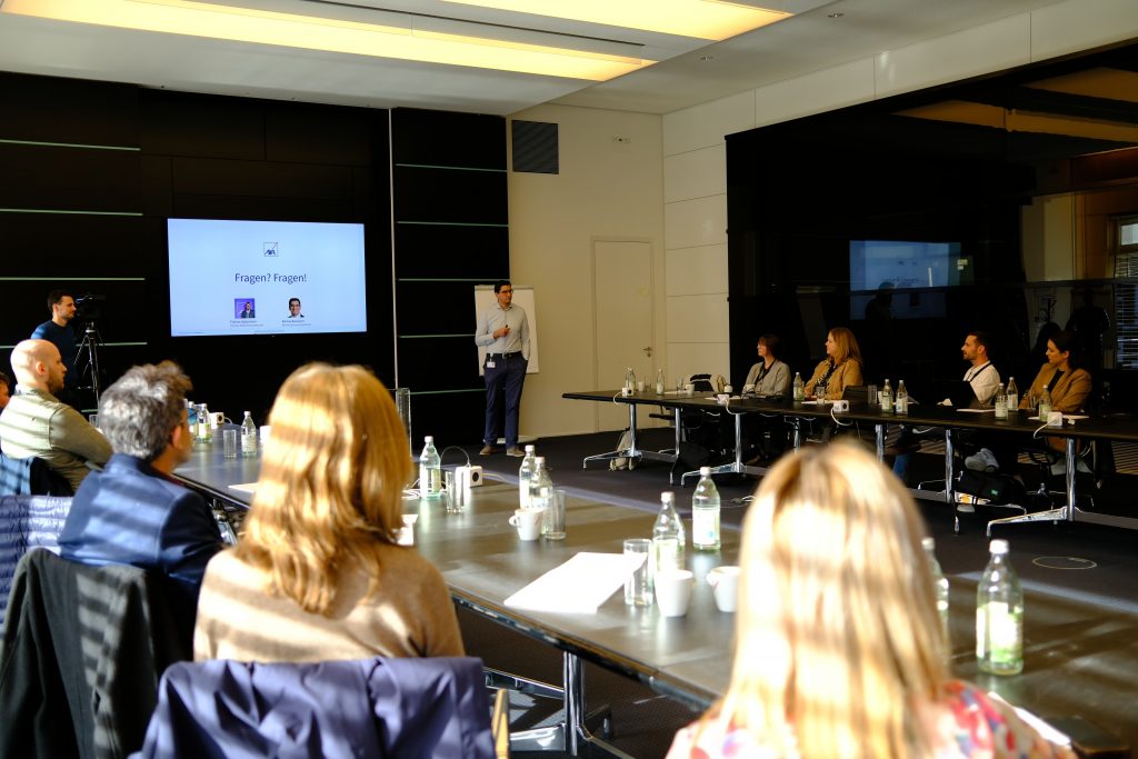 Zwei Männer halten eine Präsentation vor einer Gruppe, die an Tischen in U-Form um sie herumsitzen