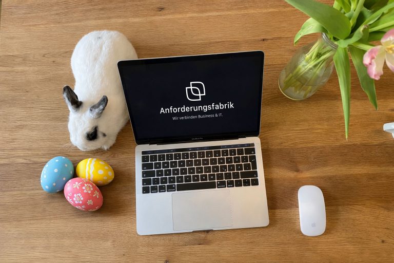 Auf einem Holztisch steht ein Laptop daneben sitzt ein schwarz-weißes Kaninchen, vor ihm drei bunte Ostereier.