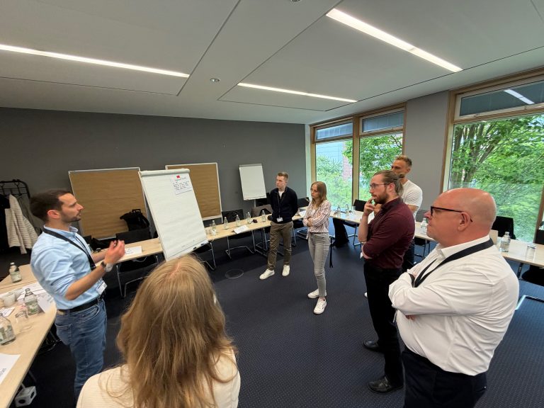 Rund 8 Personen in einem Konferenzraum stehen um ein Flipchart und diskutieren miteinander