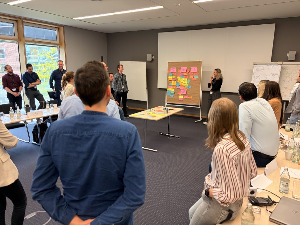 Rund 30 Personen in einem Konferenzraum blicken nach vorne: Vorne werden auf einer Metwawand Post-its mit Feedback gesammelt und moderiert